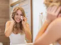 Uśmiechnięta blondynka w łazience pokazuje, jak nakładać tonik na twarz wacikiem, dbając o swoją cerę.