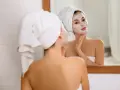 Kobieta z ręcznikiem na głowie nakłada na twarz domową maseczkę nawilżającą, podziwiając efekt w lustrze.