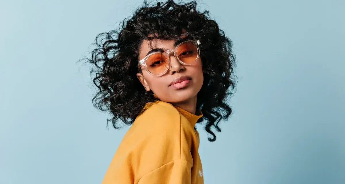 Dziewczyna z burzą loków, w okularach i żółtym swetrze, prezentuje efekt metody curly girl.