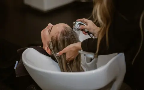 Relaks w salonie fryzjerskim: delikatne mycie włosów odżywką. Woda spływa po głowie, a dłonie fryzjerki czule pielęgnują pasma.