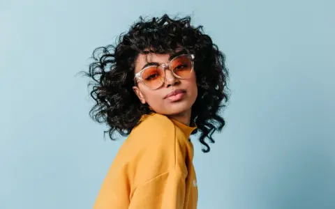 Dziewczyna z burzą loków, w okularach i żółtym swetrze, prezentuje efekt metody curly girl.