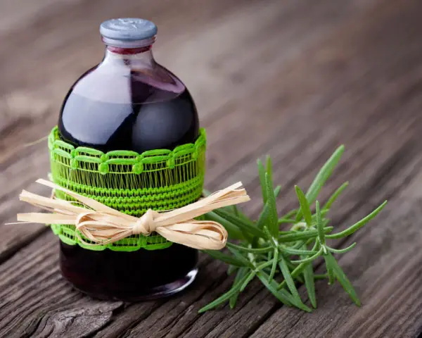Butelka z ciemnym płynem, ozdobiona zieloną koronką i słomianą kokardką, obok gałązka rozmarynu. To naturalny napar z rozmarynu na włosy.