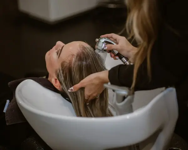 Relaks w salonie fryzjerskim: delikatne mycie włosów odżywką. Woda spływa po głowie, a dłonie fryzjerki czule pielęgnują pasma.