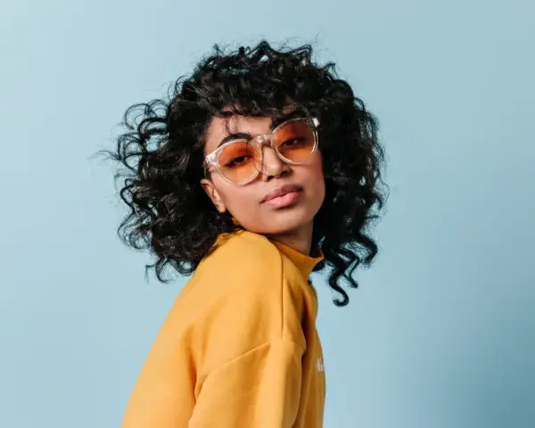 Dziewczyna z burzą loków, w okularach i żółtym swetrze, prezentuje efekt metody curly girl.