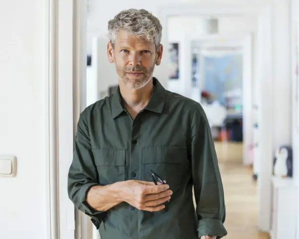 Eleganckie fryzury męskie po 50-tce: siwy, krótko ścięty włos i zarost dodają mu charyzmy. Mężczyzna w zielonej koszuli stoi w korytarzu.