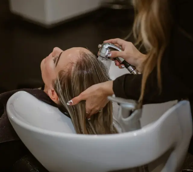 Relaks w salonie fryzjerskim: delikatne mycie włosów odżywką. Woda spływa po głowie, a dłonie fryzjerki czule pielęgnują pasma.