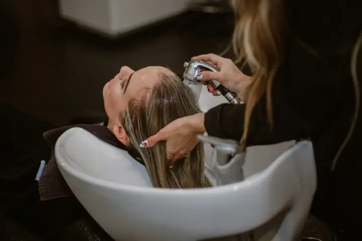 Relaks w salonie fryzjerskim: delikatne mycie włosów odżywką. Woda spływa po głowie, a dłonie fryzjerki czule pielęgnują pasma.
