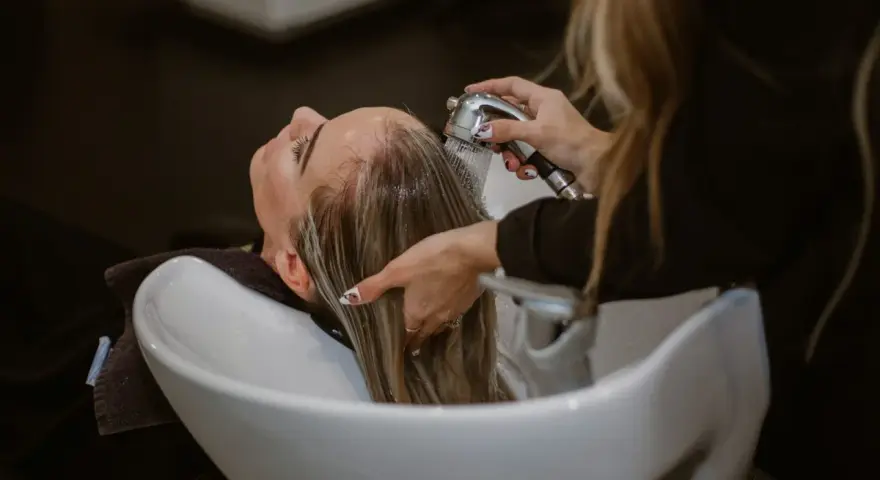 Relaks w salonie fryzjerskim: delikatne mycie włosów odżywką. Woda spływa po głowie, a dłonie fryzjerki czule pielęgnują pasma.