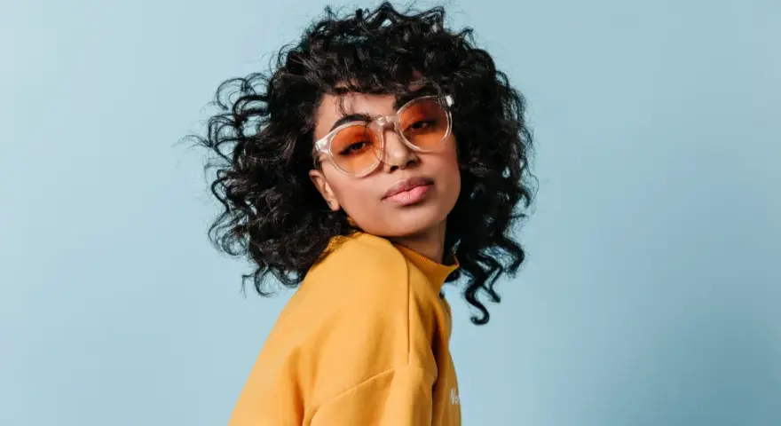 Dziewczyna z burzą loków, w okularach i żółtym swetrze, prezentuje efekt metody curly girl.