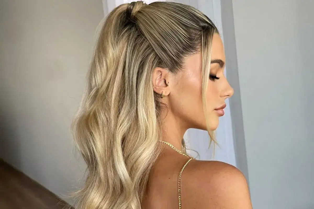 Eleganckie fryzury na wesele jako gość: blond włosy upięte w wysoki kucyk z luźnymi falami i kilkoma pasmami opadającymi na twarz.