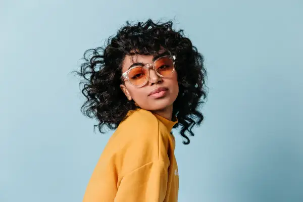 Dziewczyna z burzą loków, w okularach i żółtym swetrze, prezentuje efekt metody curly girl.